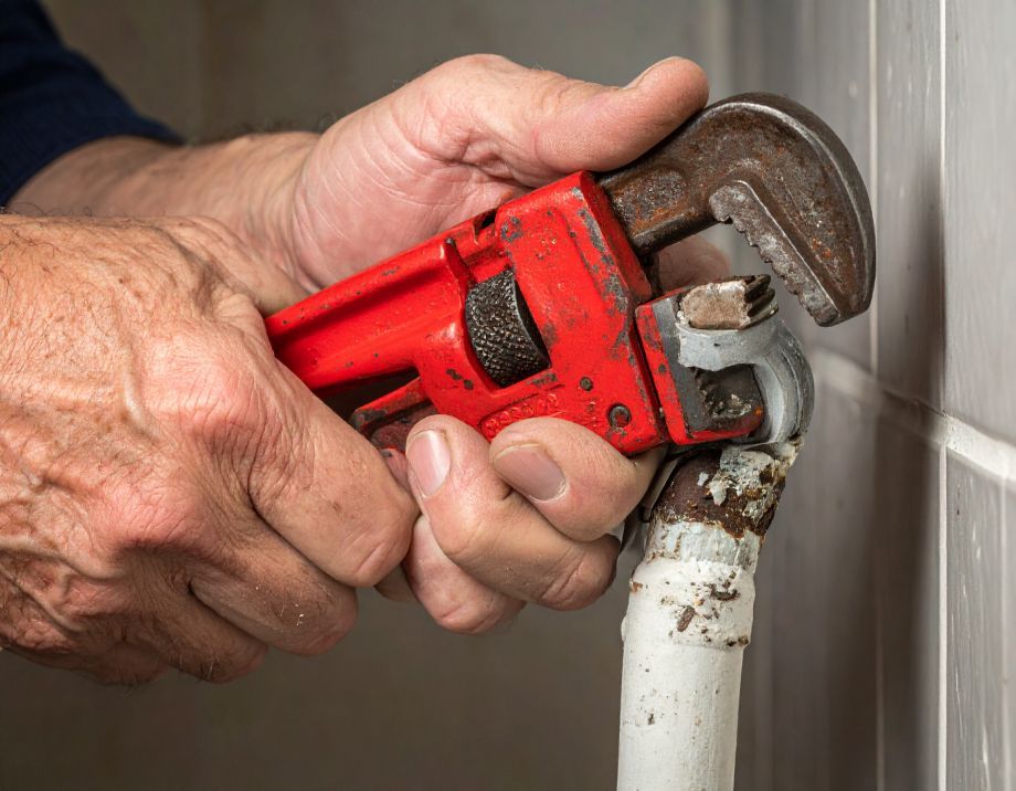 Hands using a red wrench to tighten a rusty pipe.