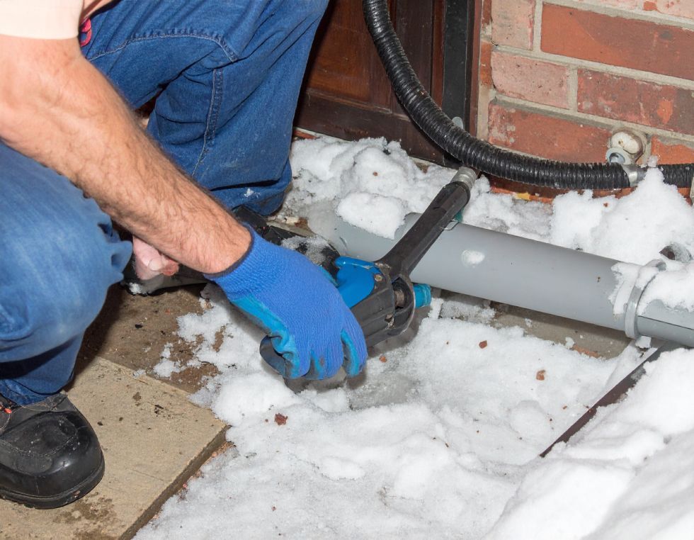 Person in blue gloves and jeans using a tool on a gray pipe outside on a snowy day.