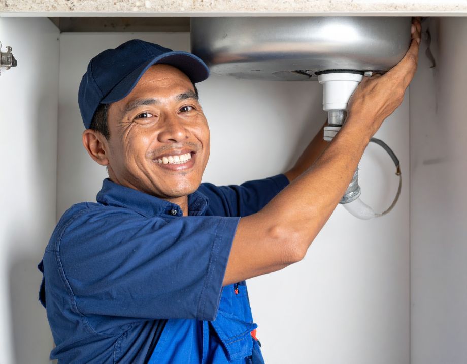 Plumber in blue uniform smiling, working under a kitchen sink.