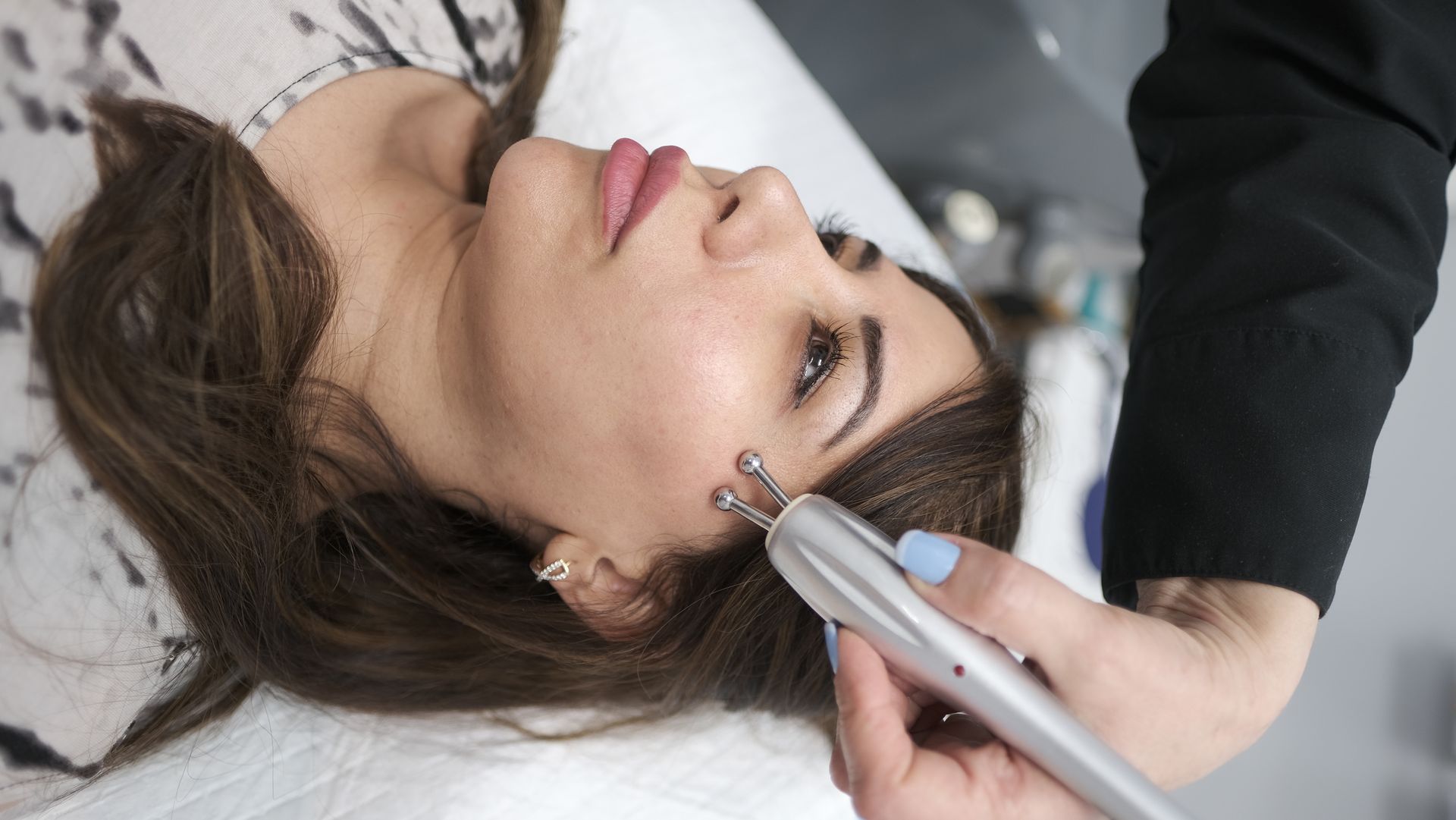 Woman receiving facial treatment with a device held by a person's hand in a medical setting.