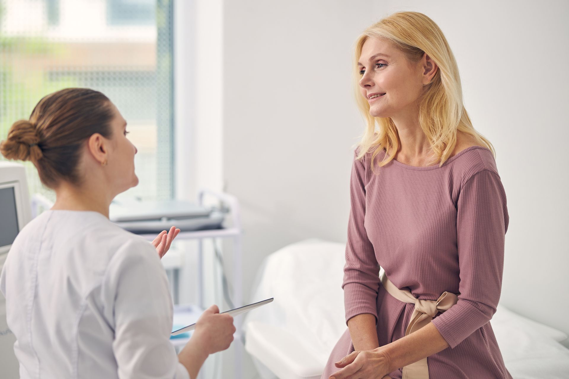 Woman in mauve dress conversing with person in white coat in medical office, possibly patient and doctor.