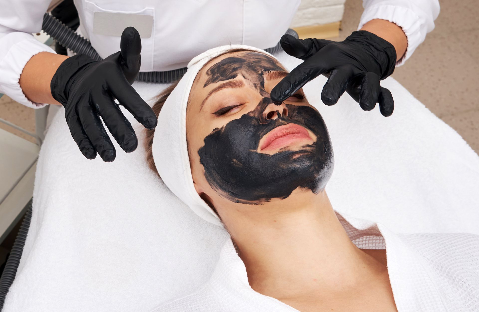 Woman with a black facial mask receiving spa treatment; therapist's hands in black gloves.