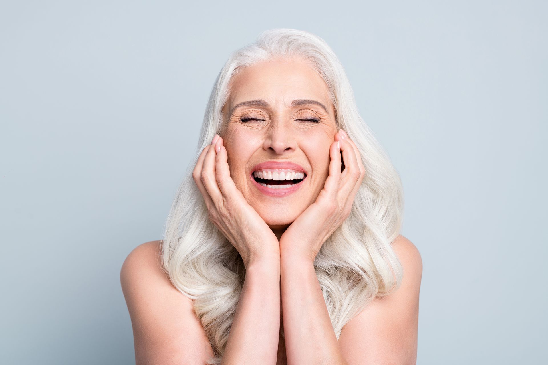 A woman with gray hair is smiling with her eyes closed and her hands on her face.