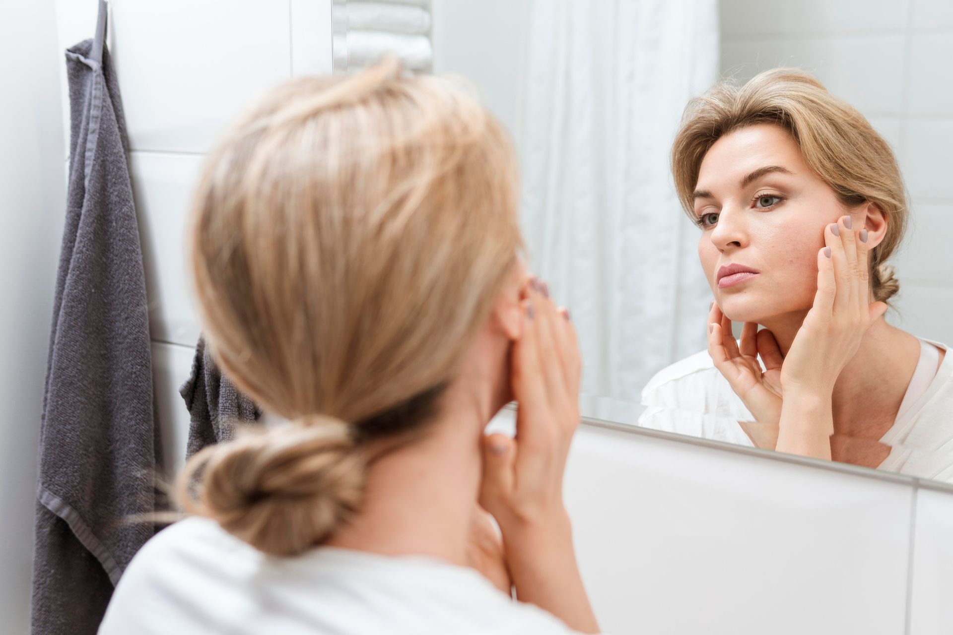 Woman looking at herself in a bathroom mirror, touching her face. White wall and a towel on the side.
