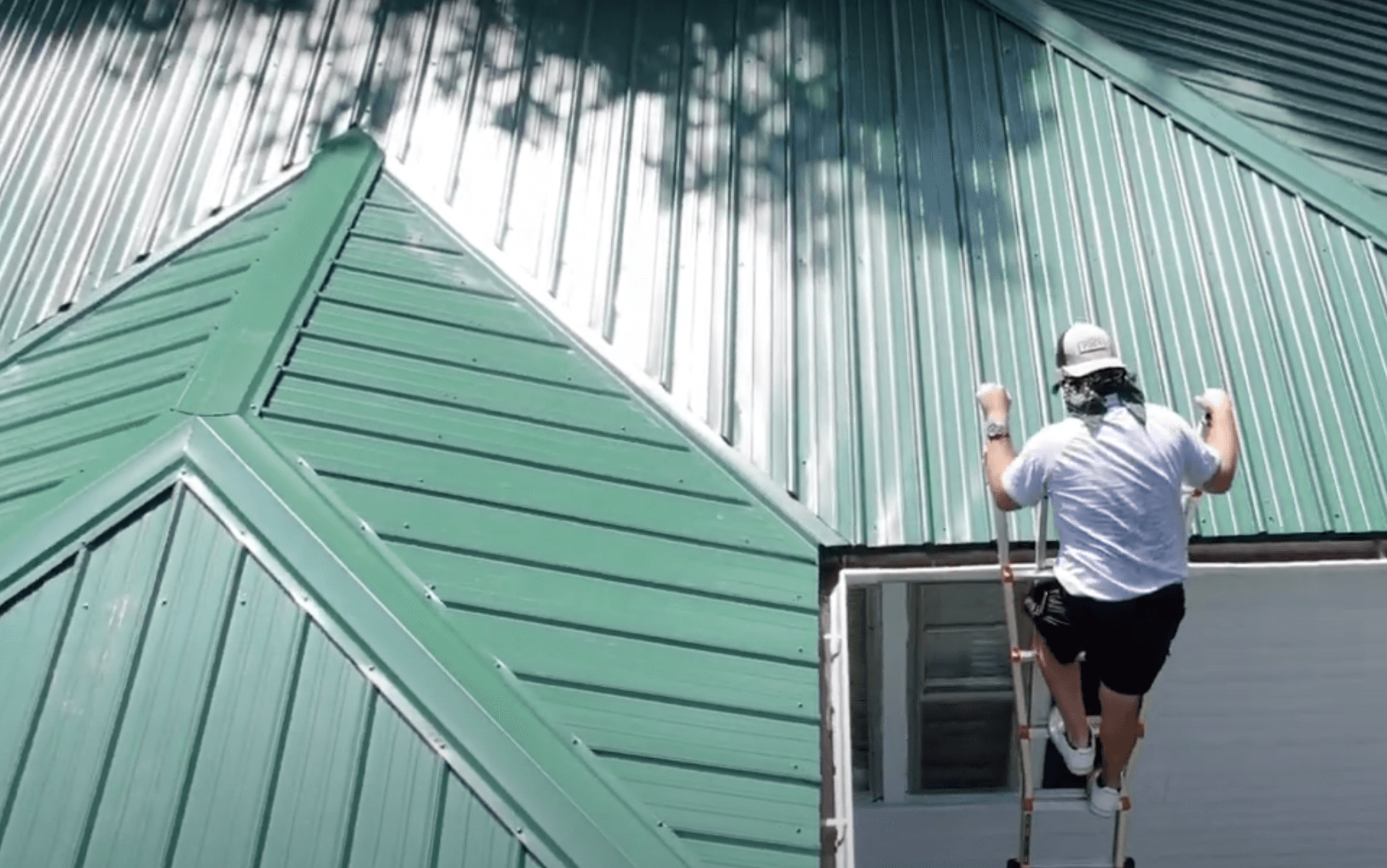 A man is standing on a ladder painting a green roof.