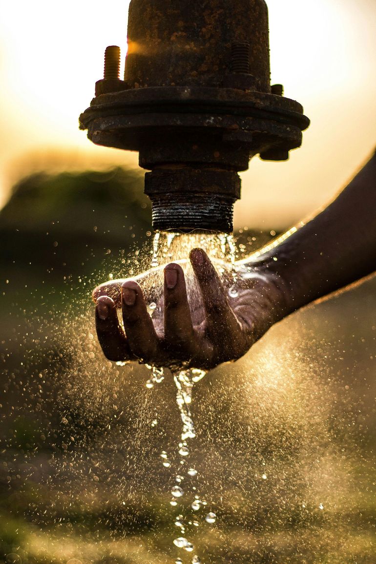 Hand cupped under flowing water from a rusty spigot, illuminated by golden sunlight.