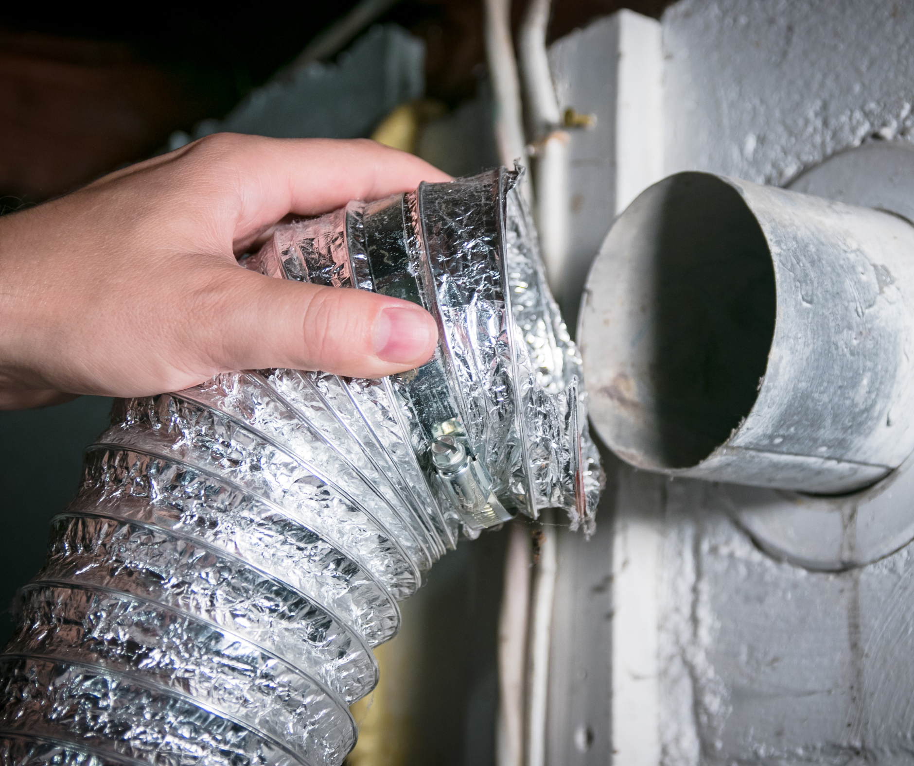 Hand holding a flexible dryer vent hose next to a metal wall outlet.