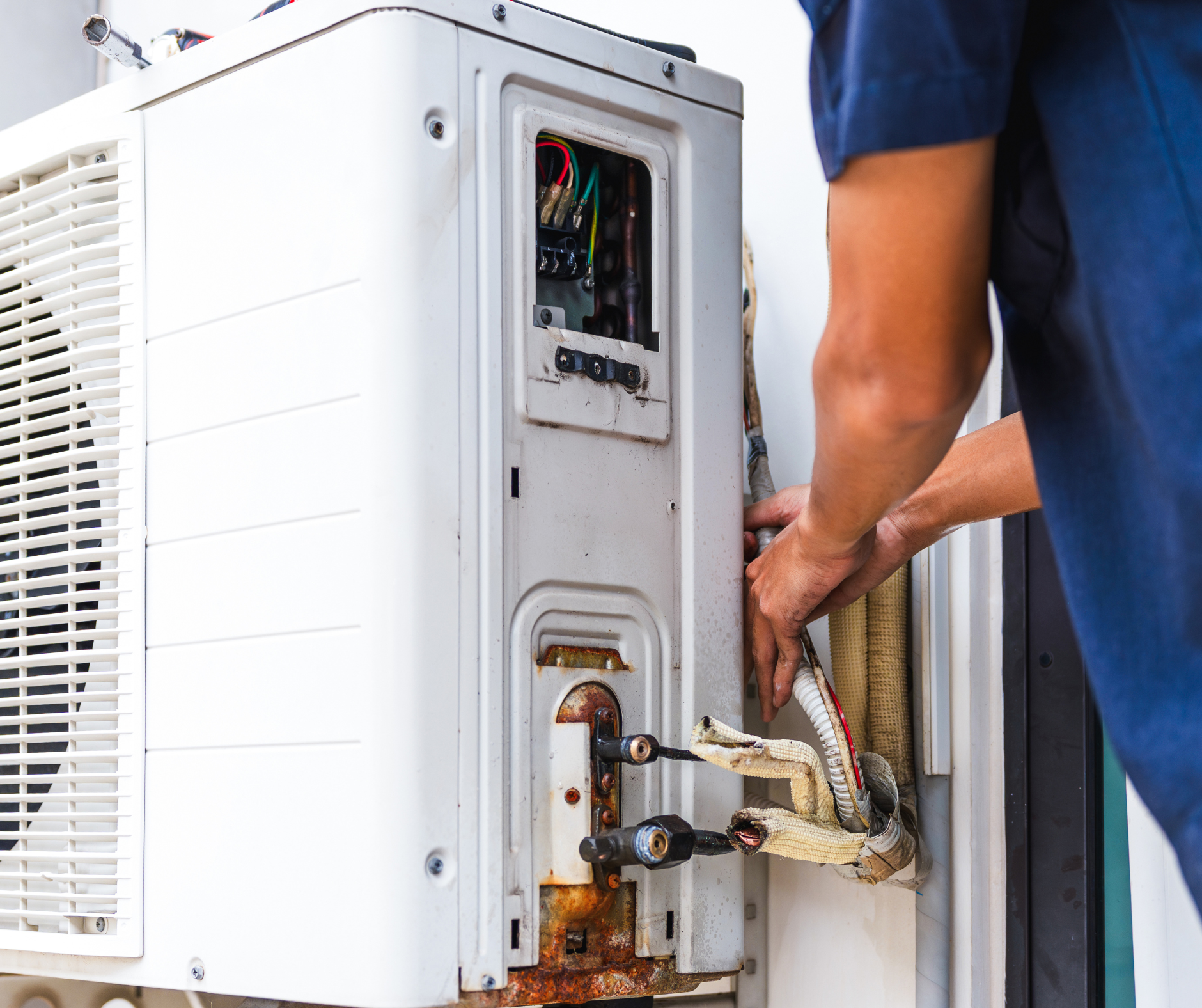 A person repairs an outdoor air conditioning unit, near a wall.