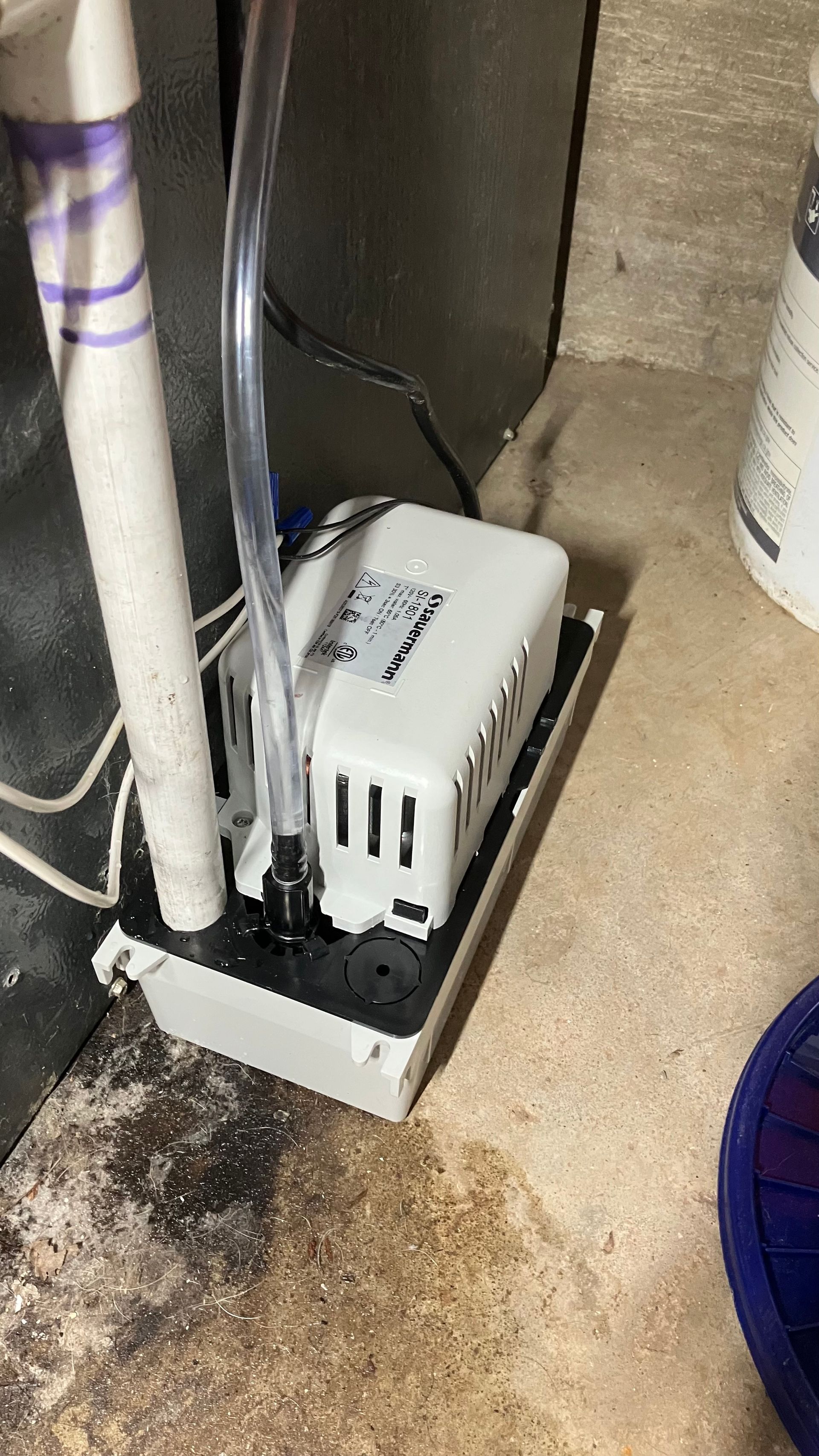 White sump pump in a basement, connected to a PVC pipe and a clear hose, standing on a wet concrete floor.