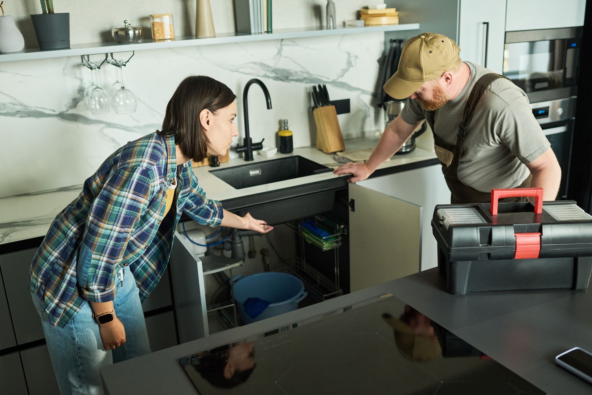 Woman explains plumbing issue to plumber repairing kitchen sink with toolbox on counter. Woman explains plumbing issue to plumber repairing kitchen sink with toolbox on counter.