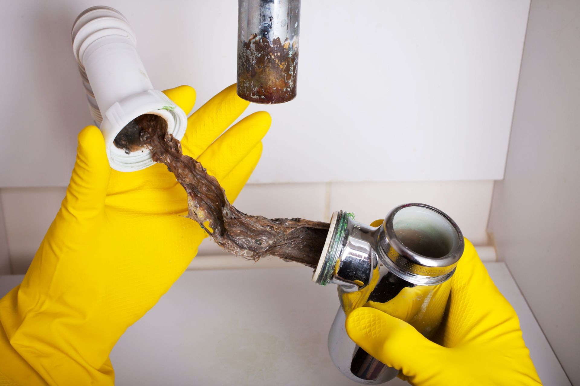 Yellow-gloved hands disconnecting a clogged white pipe under a sink, with dirty water and debris spilling out