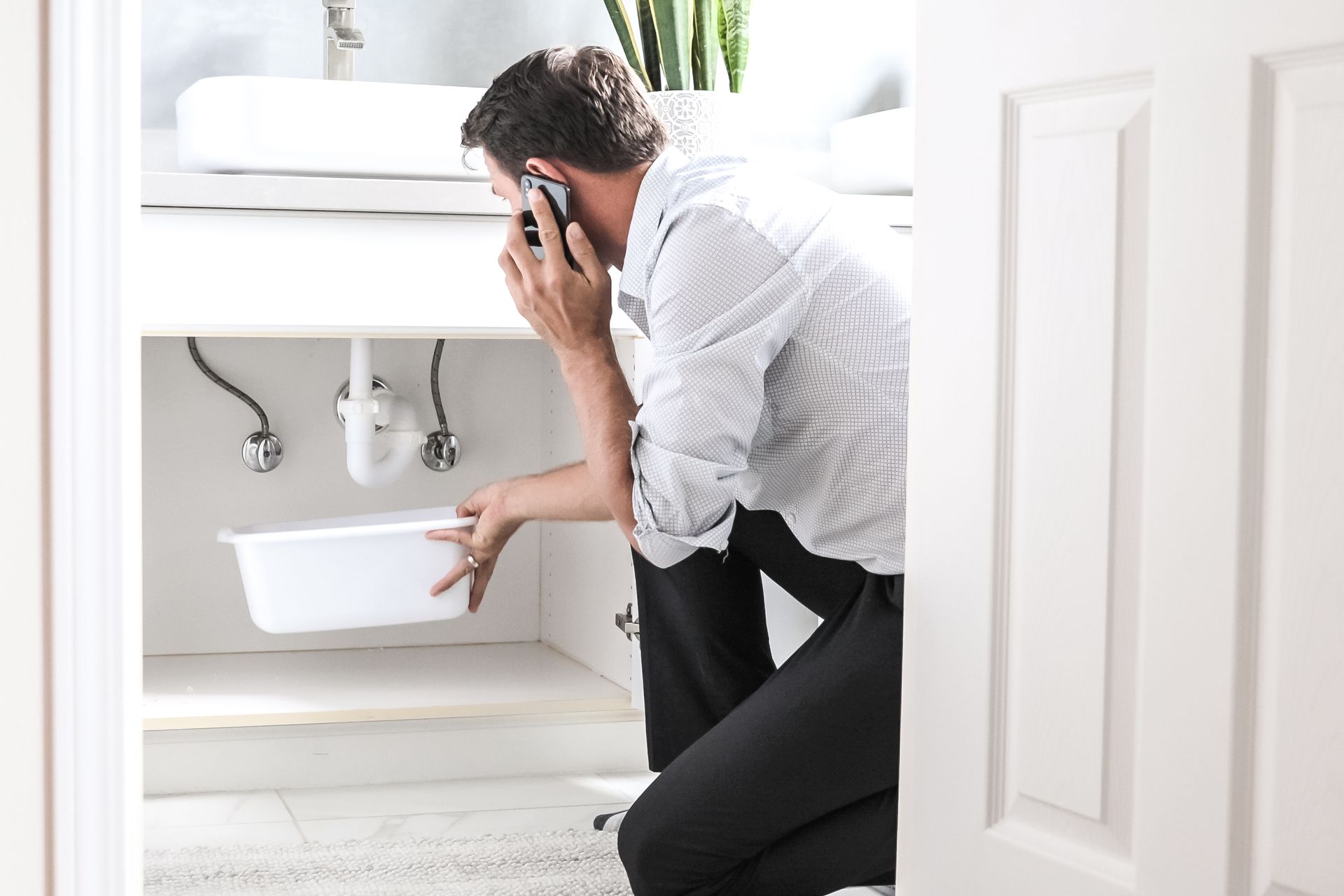 Man crouching under a sink while talking on a phone, holding a white basin in a bathroom.
