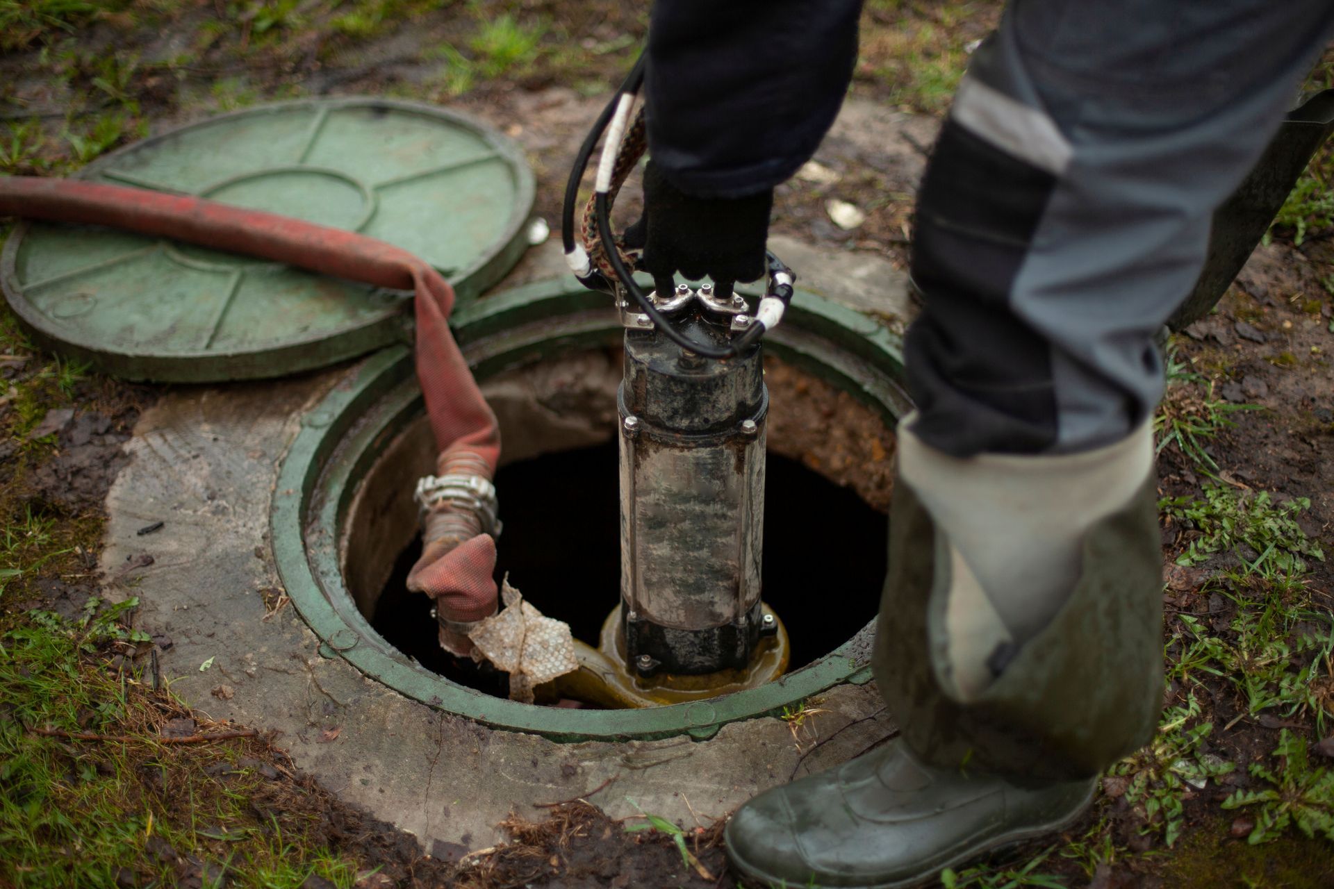 Workers opening a green manhole cover with a hose inserted into the sewer opening