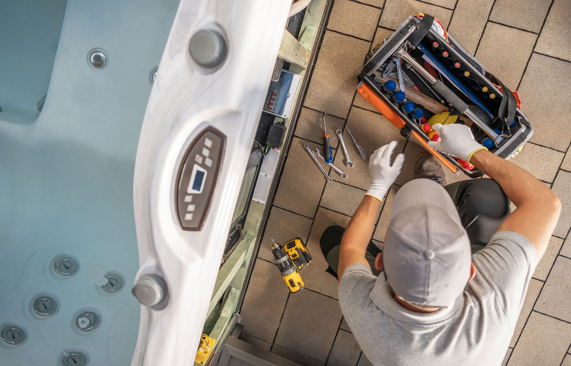 Technician repairing equipment beside a hot tub, with tools and wiring on a tiled patio.