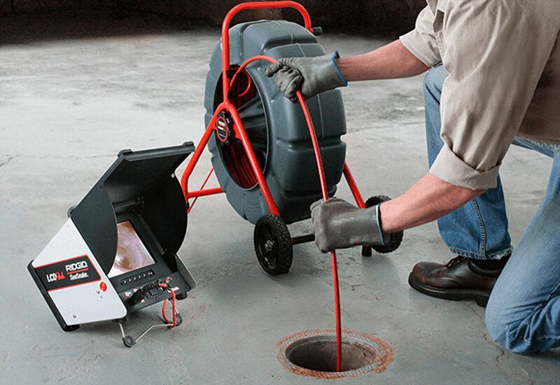 Worker using a red drain cleaning machine to feed cable into a floor drain in a concrete room
