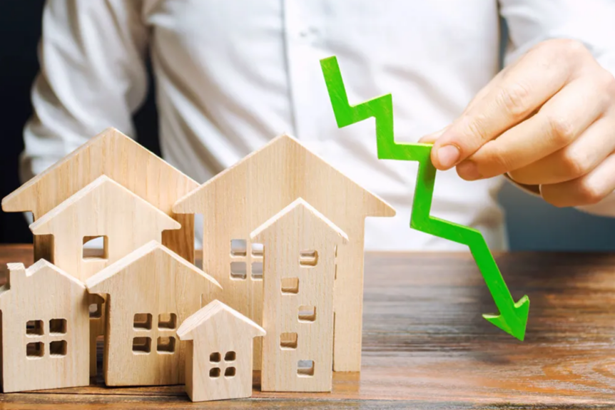 Person holding a downward-pointing green arrow next to wooden houses, representing a decline in the real estate market.