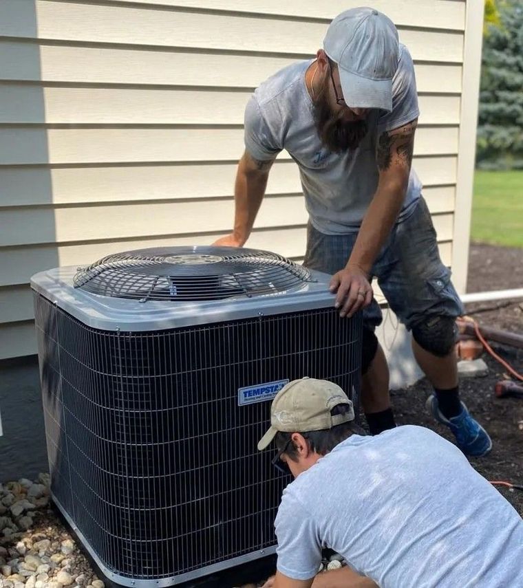 Two workers installing an air conditioner unit outside a home. One kneels, the other stands.