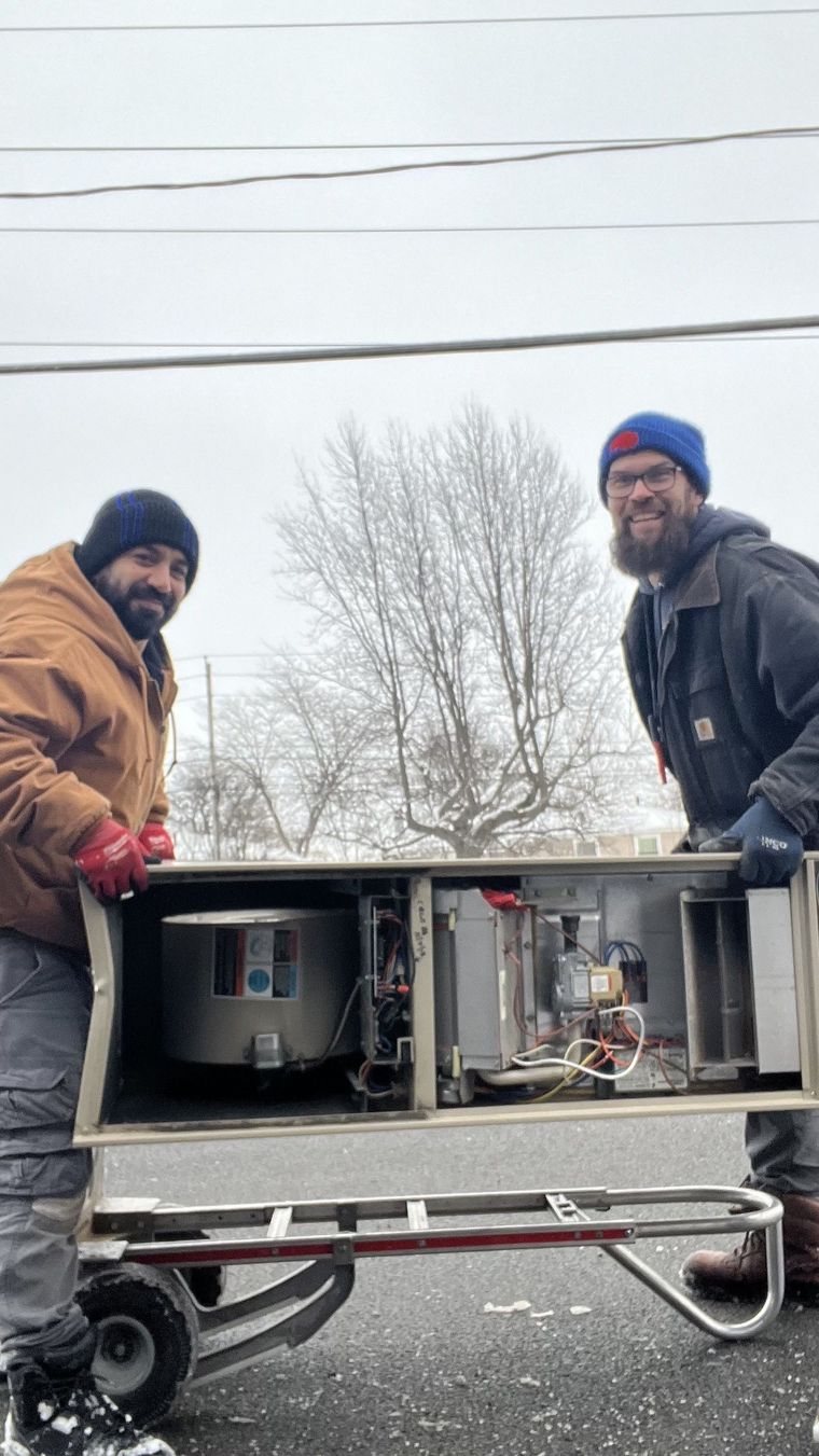Two people moving a furnace on a hand truck outside in snowy conditions.