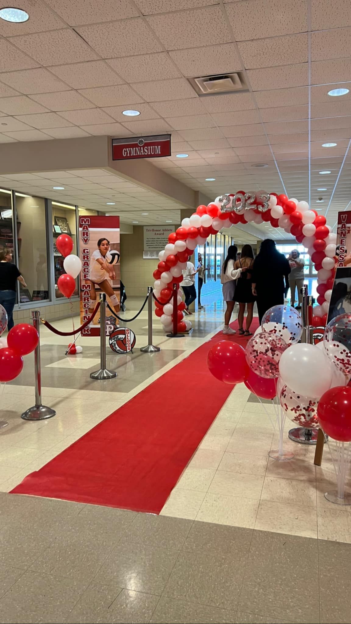 Crown Point High School's Volleyball sports award ceremony balloon decor designed to create an unforgettable entrance. Featuring a spiral red and white classic balloon arch topped with '2023' foil numbers, balloon bouquets lining the red carpet, and signature confetti balloon accents for a truly exciting and memorable night.