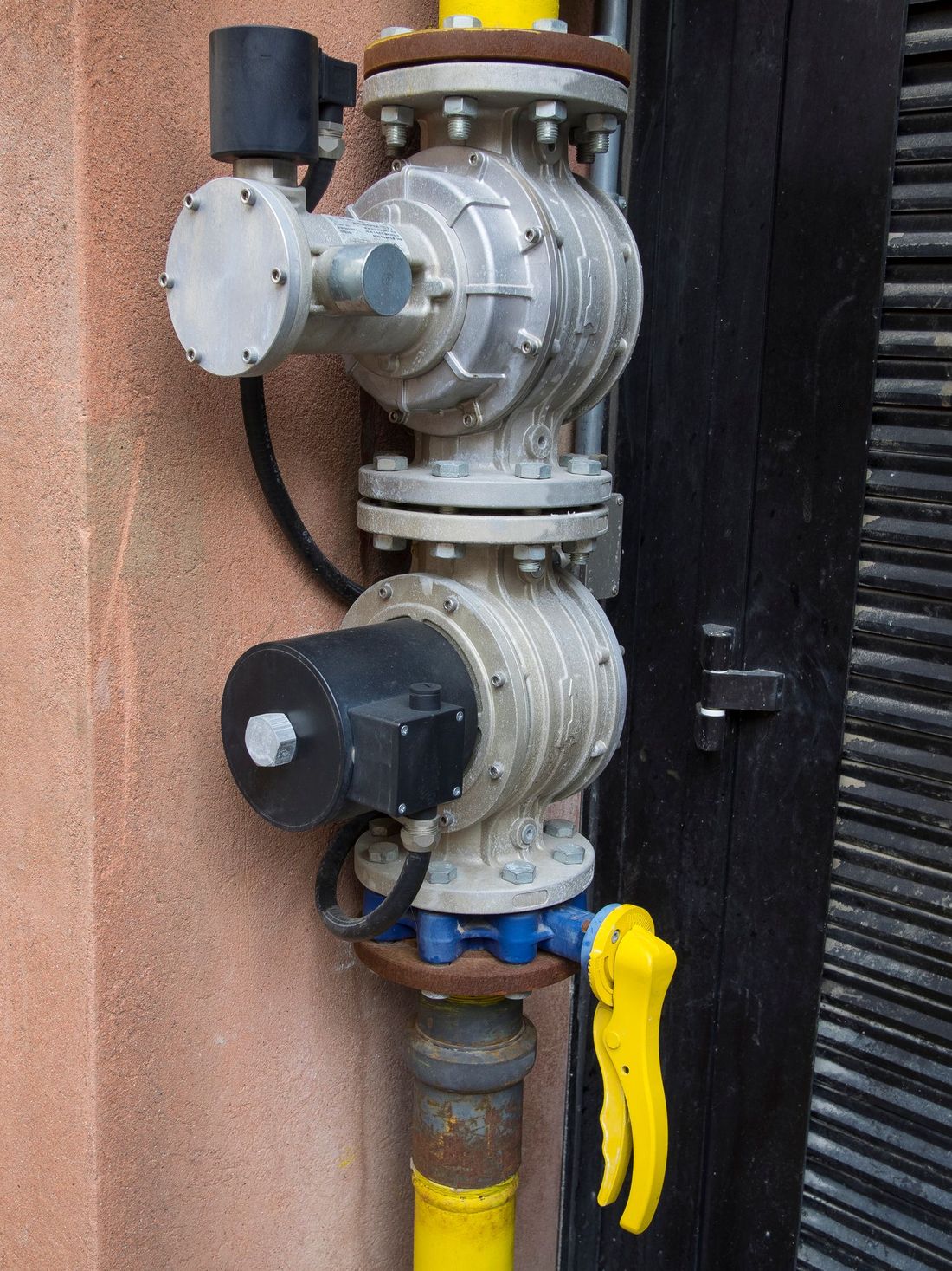 Due valvole del gas industriali collegate a un tubo giallo, montate su un edificio, con maniglia gialla.