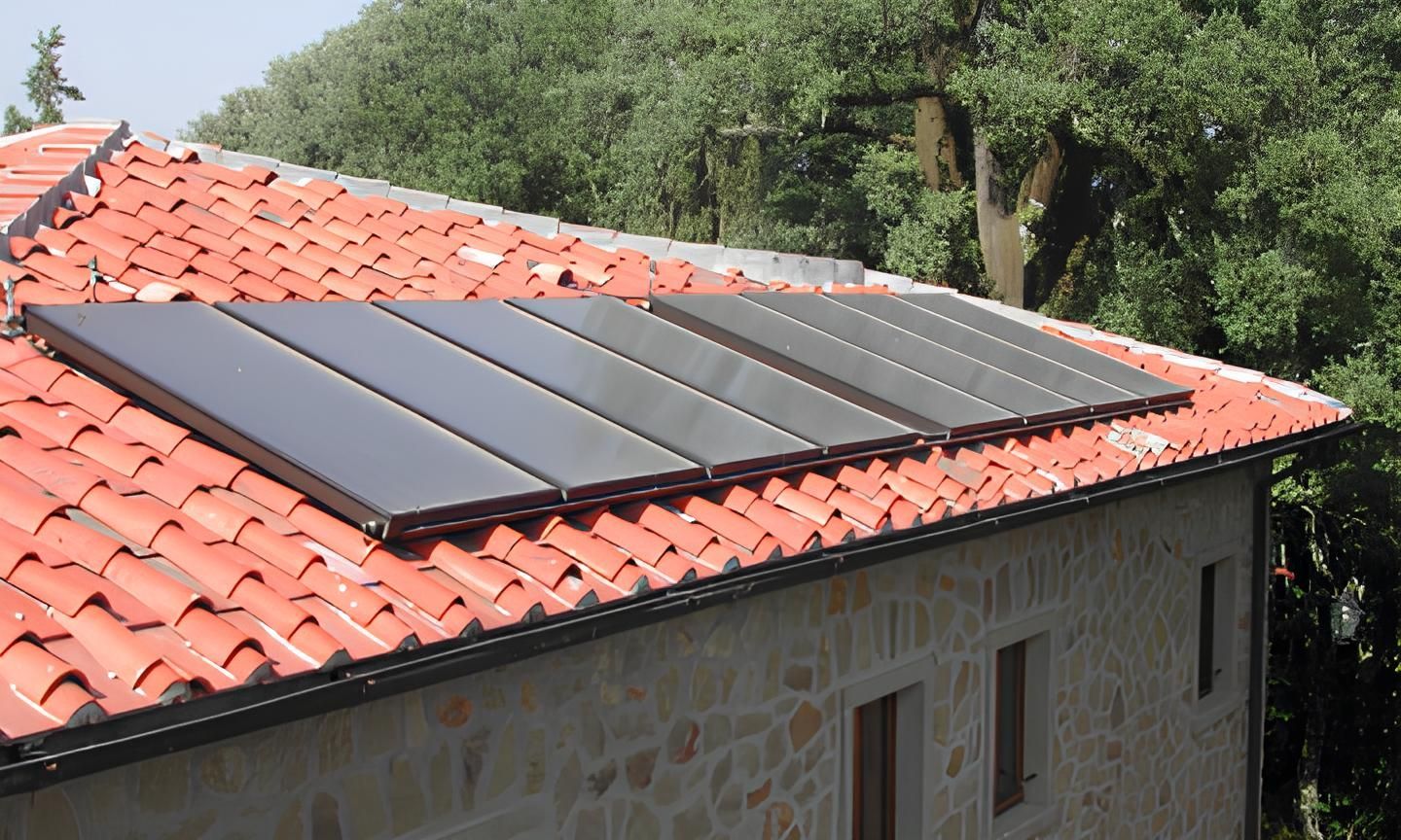 Pannelli solari sul tetto di tegole rosse di un edificio in pietra, con alberi sullo sfondo.