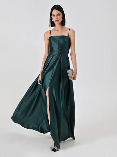 Donna in abito verde smeraldo con spacco profondo che tiene in mano una pochette, in piedi in uno studio.