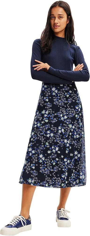 Donna con top blu navy e gonna floreale, in piedi con le braccia incrociate, che indossa scarpe da ginnastica.