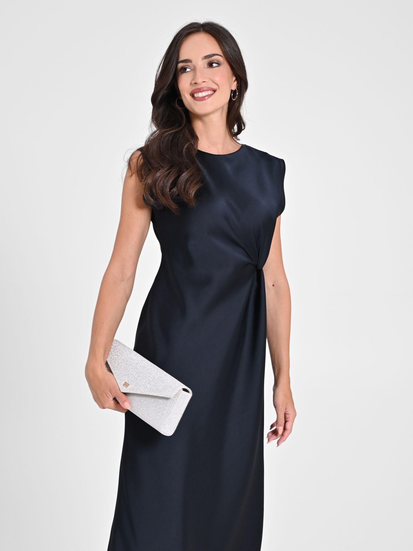 Donna in abito blu scuro che tiene in mano una pochette bianca, sorridente.