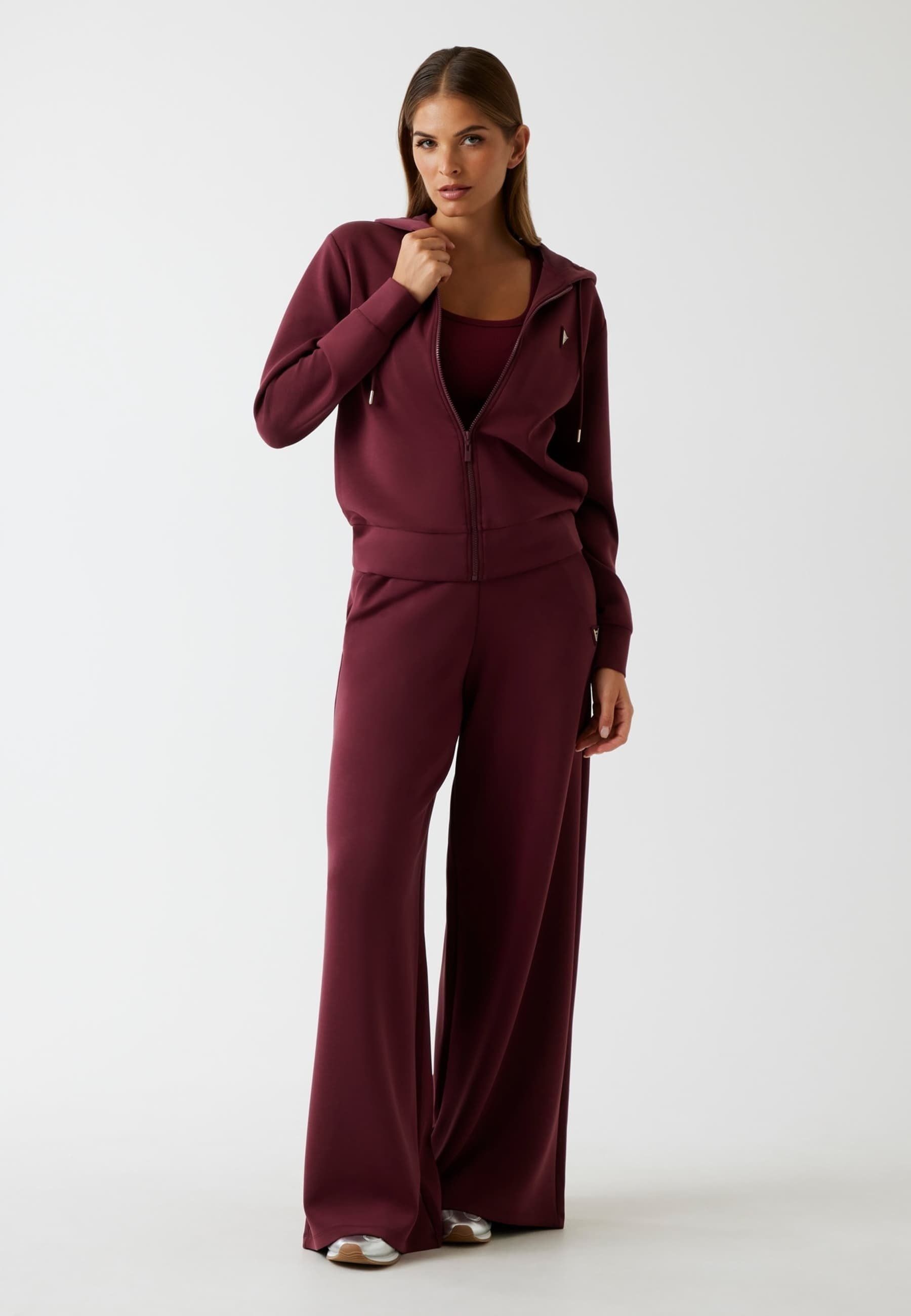 Donna in tuta color bordeaux, in piedi, che si tocca il collo. Sfondo chiaro.