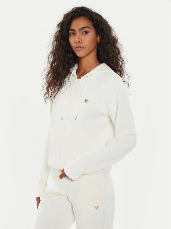 Donna con felpa bianca e pantaloni della tuta. È in piedi, guarda la telecamera. Espressione neutra.