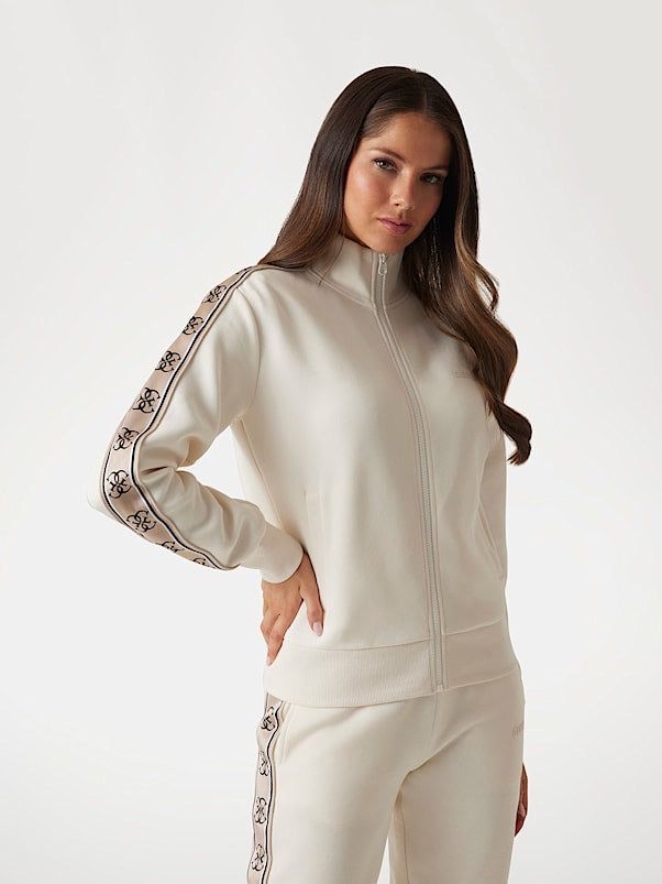 Donna in tuta color crema, in piedi, mano sul fianco. Giacca con logo e cerniera. Sfondo bianco.