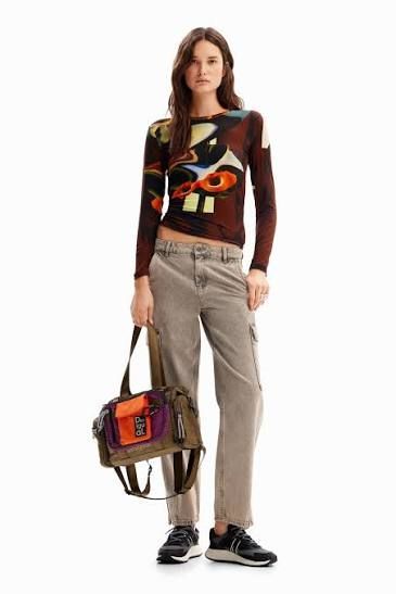 Donna con top colorato e pantaloni color cuoio, con in mano una borsa e delle scarpe da ginnastica.