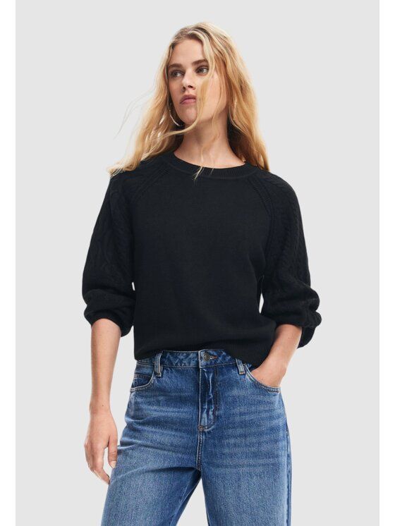 Donna che indossa un maglione nero e jeans blu, in piedi in casa con le mani in tasca.