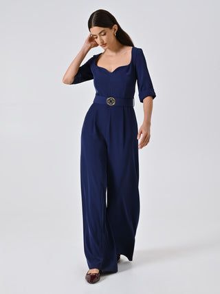 Donna in tuta blu navy con scollatura smerlata e cintura, in piedi su sfondo bianco.