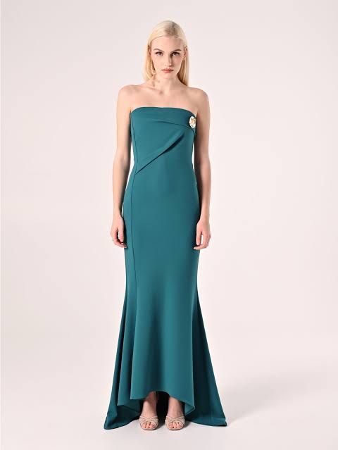 Donna in abito senza spalline color verde acqua con spilla, in piedi davanti a uno sfondo bianco.