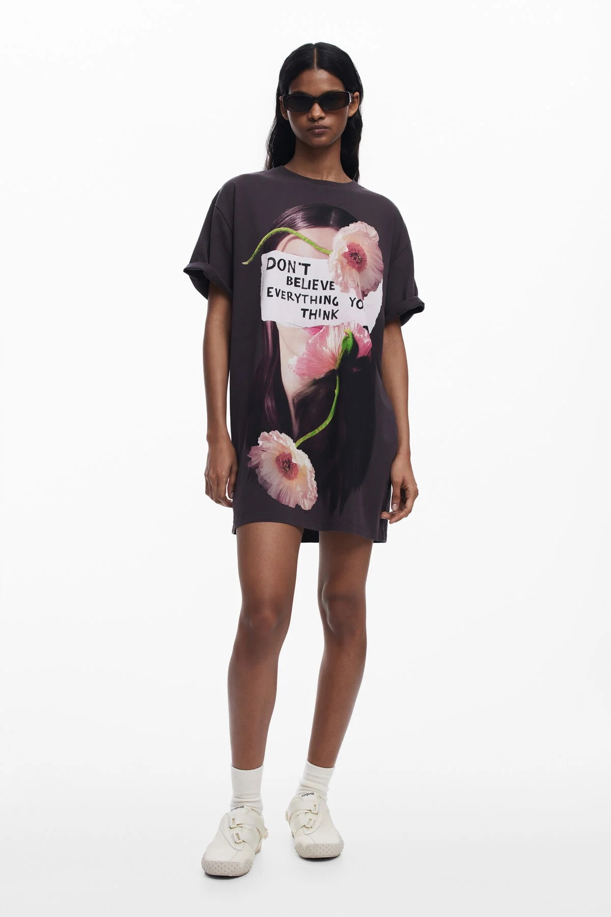 Donna che indossa un abito-t-shirt scuro con fiori e scritte, maniche arrotolate, occhiali da sole e scarpe da ginnastica bianche.