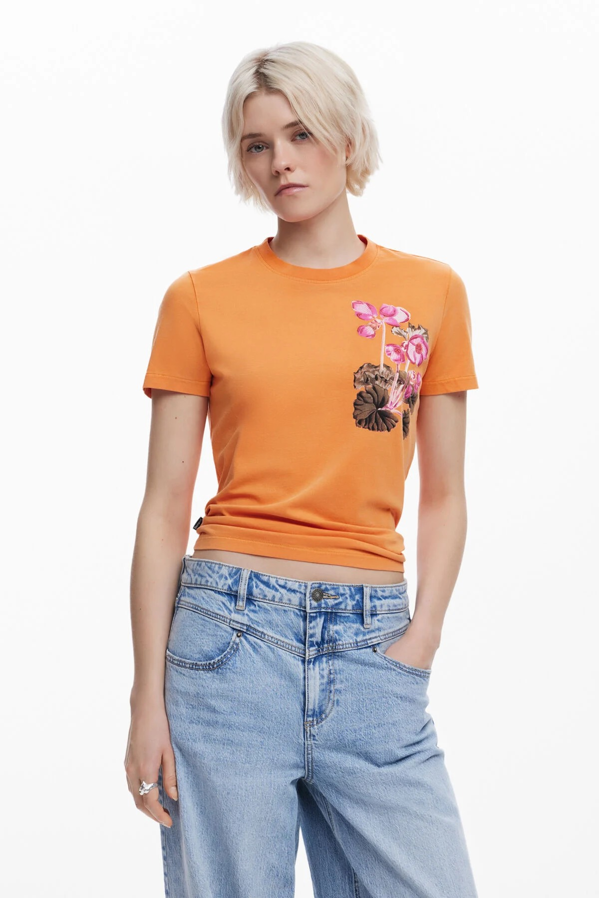 Donna con camicia arancione e jeans, mani in tasca; la camicia ha una stampa floreale.