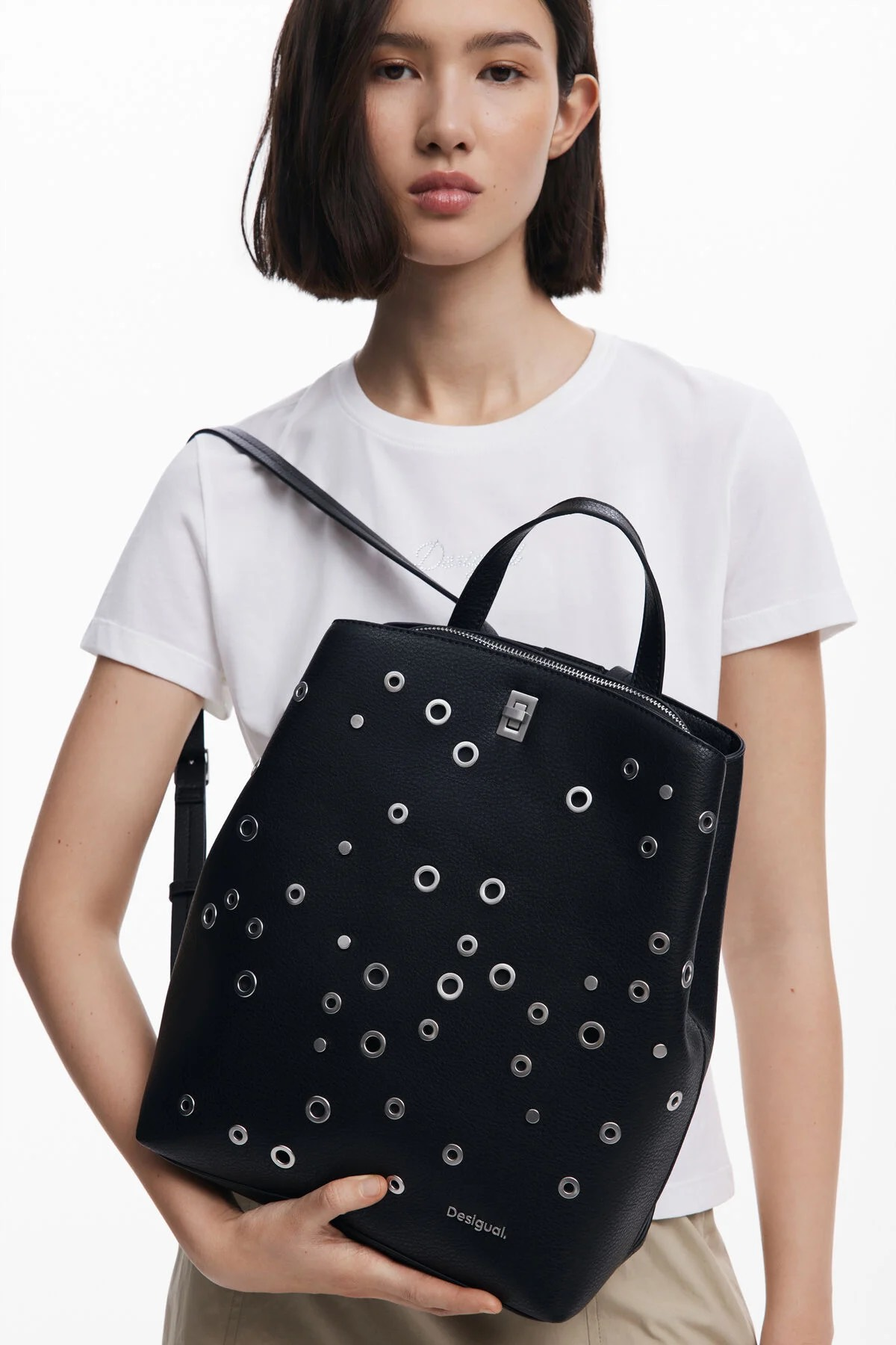 Donna con in mano uno zaino nero con occhielli argentati. Indossa una camicia bianca e pantaloni color cachi.