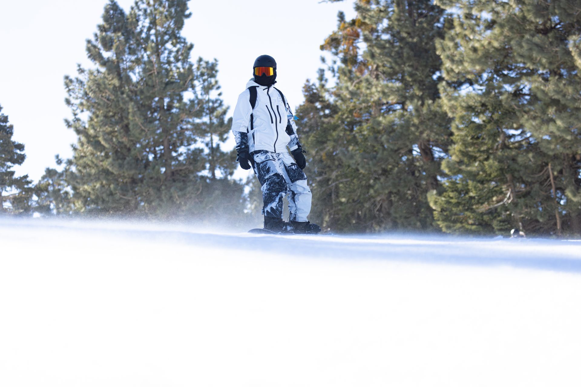 Snowboarder in white outfit on a snowy slope, trees in background, sunny day.