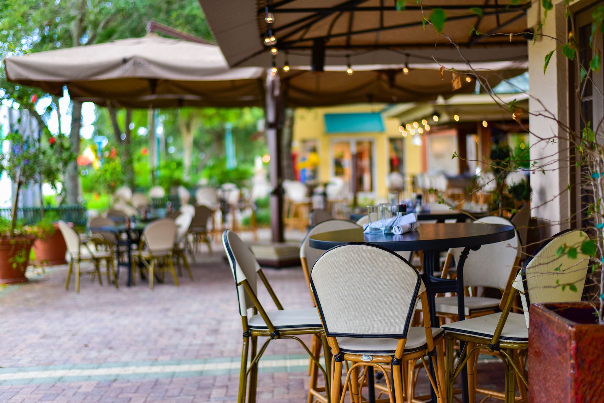 Outdoor cafe seating area featuring round dark tables, cream-colored chairs, and large tan umbrellas on a brick patio.