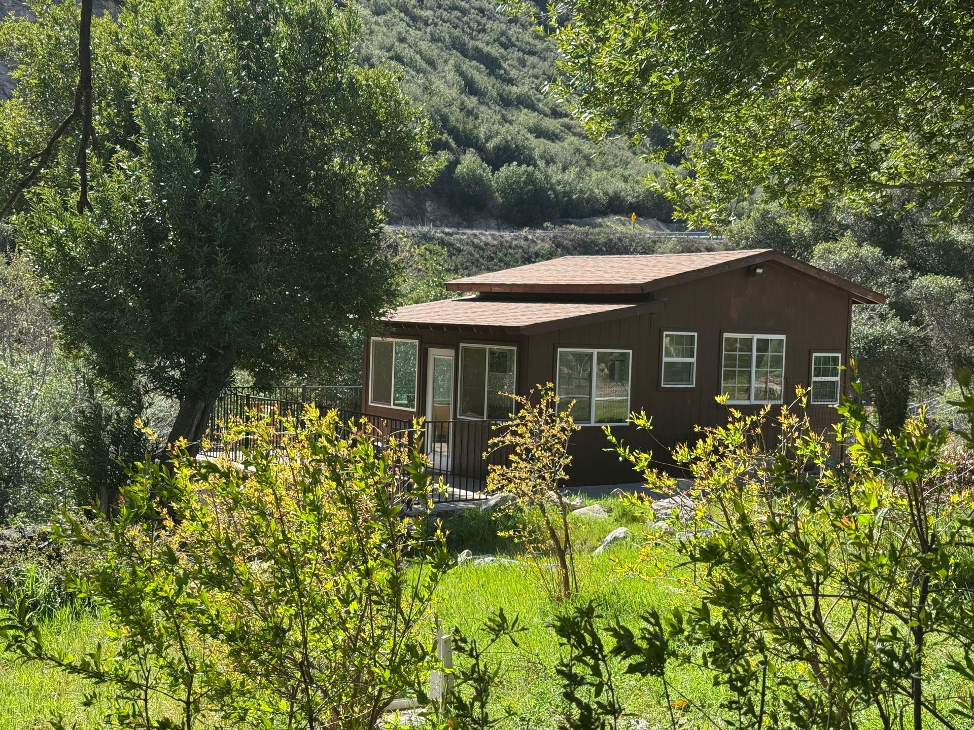 A small, brown wooden cabin nestled in a lush, green, sunlit landscape with trees and bushes in the foreground.