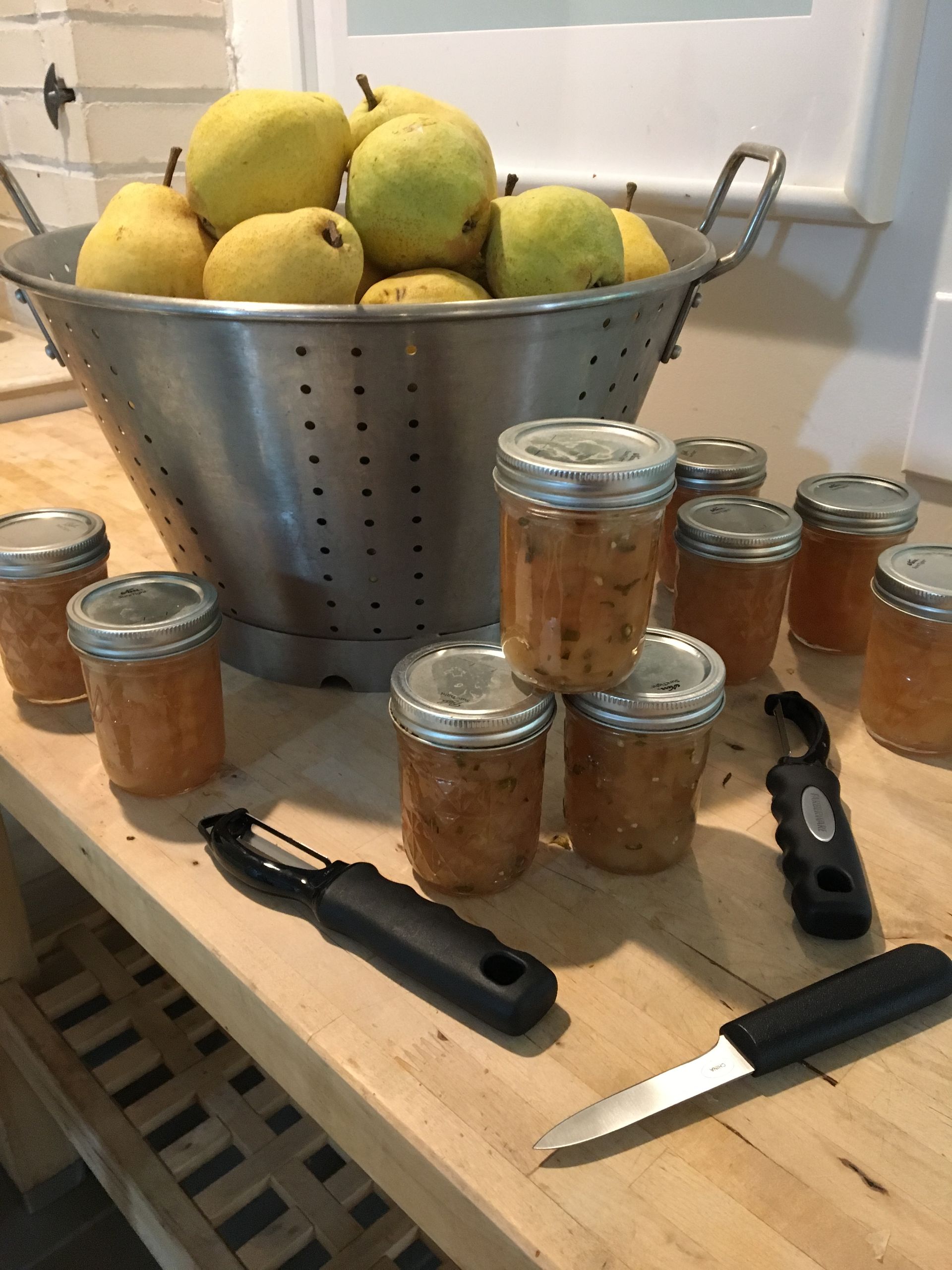A colander filled with yellow pears sits on a counter next to several glass jars of preserves, a peeler, and a knife.