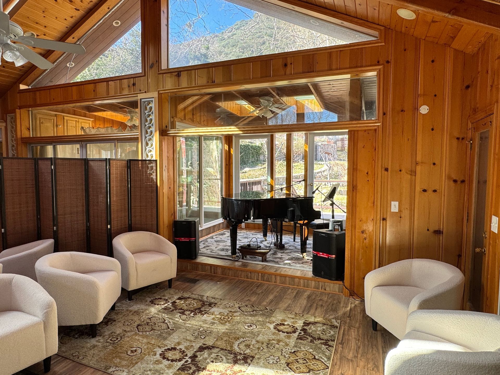 A wood-paneled room with four beige armchairs arranged around a patterned rug, facing a grand piano on a raised platform.