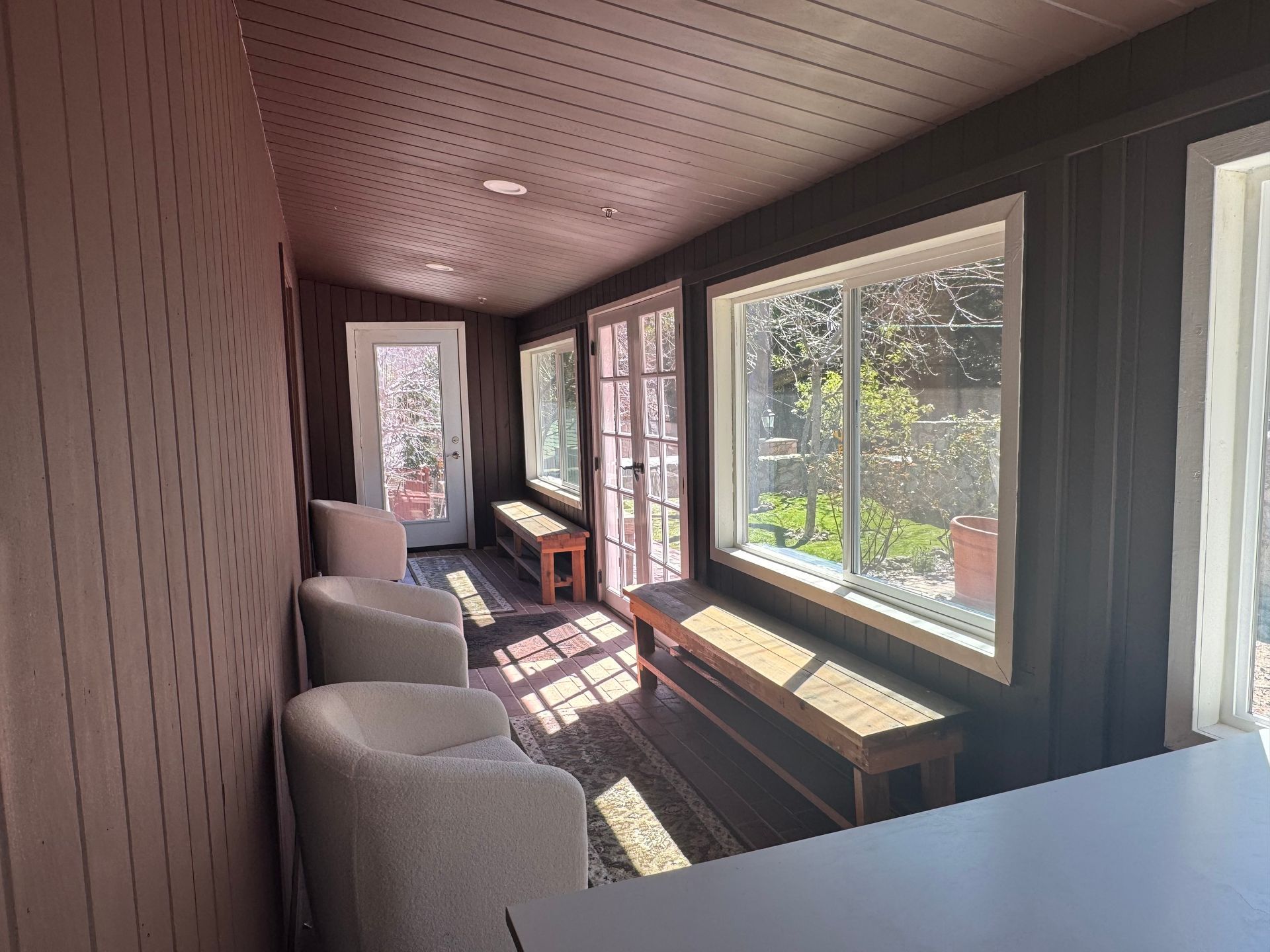 A sunroom with dark brown walls, a patterned ceiling, three light-colored armchairs, two wooden benches, and large windows.