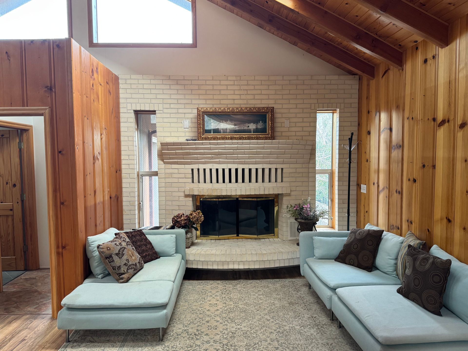 A bright living room with wood paneled walls, a large white brick fireplace, and two light blue sofas facing each other.