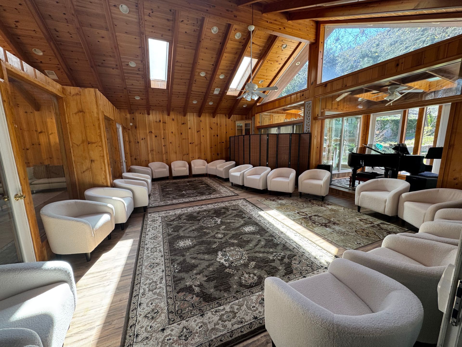 A spacious, wood-paneled room with vaulted ceilings and skylights, arranged with many light-colored armchairs in a circle.