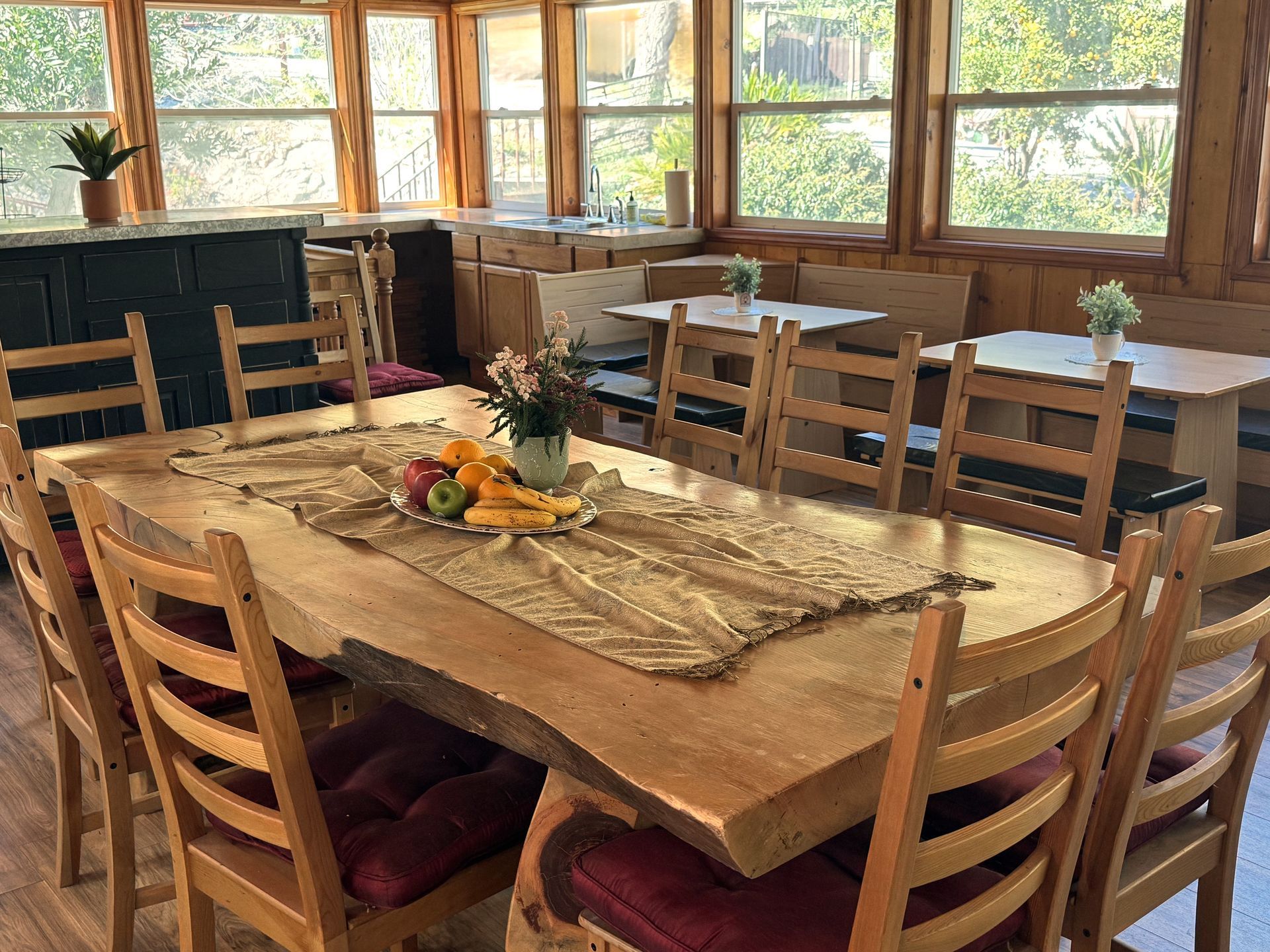 A rustic dining room with a long wooden table, multiple chairs, and windows overlooking a green outdoor space.