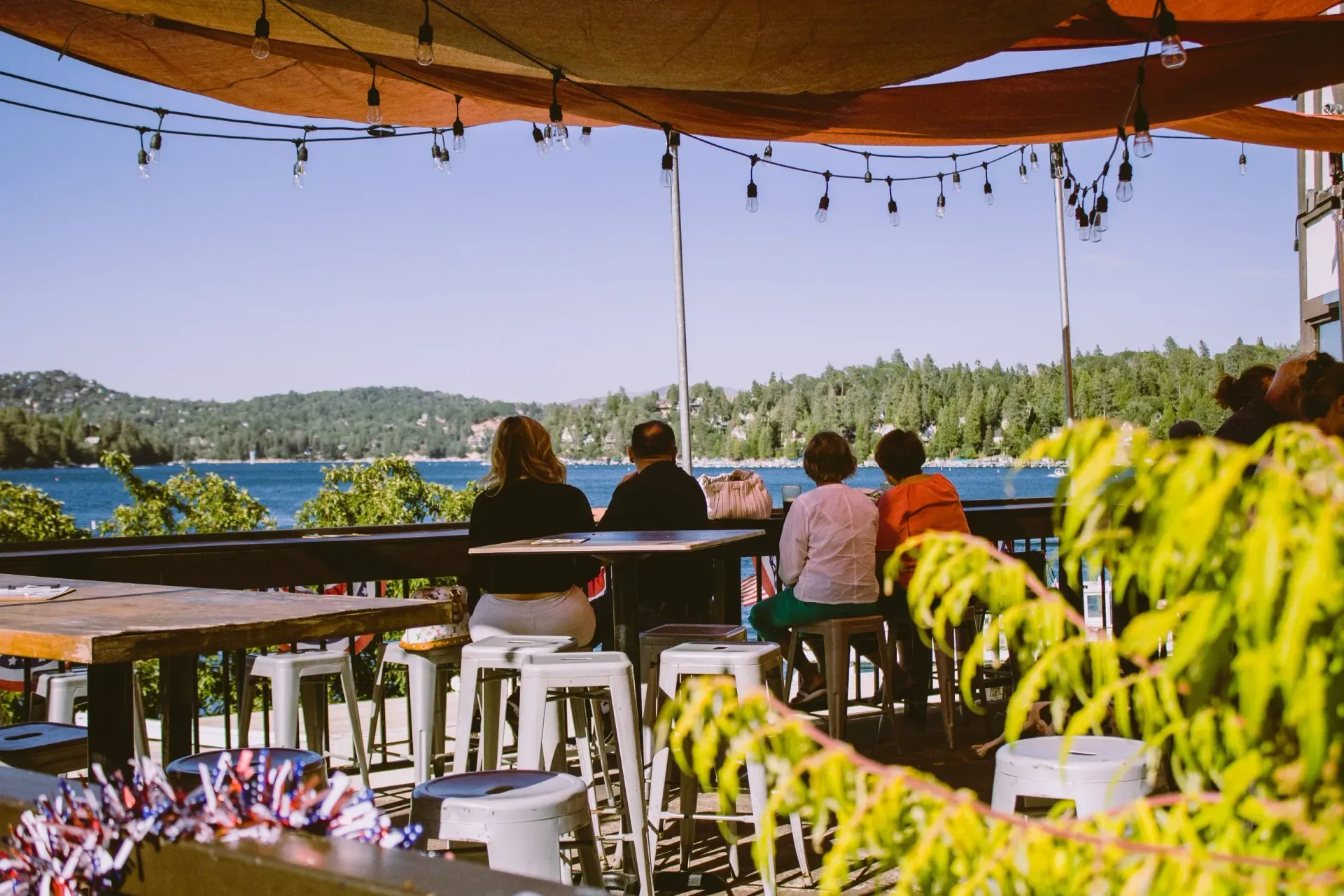 People seated at a scenic outdoor bar overlooking a lake surrounded by trees under a string-light canopy.
