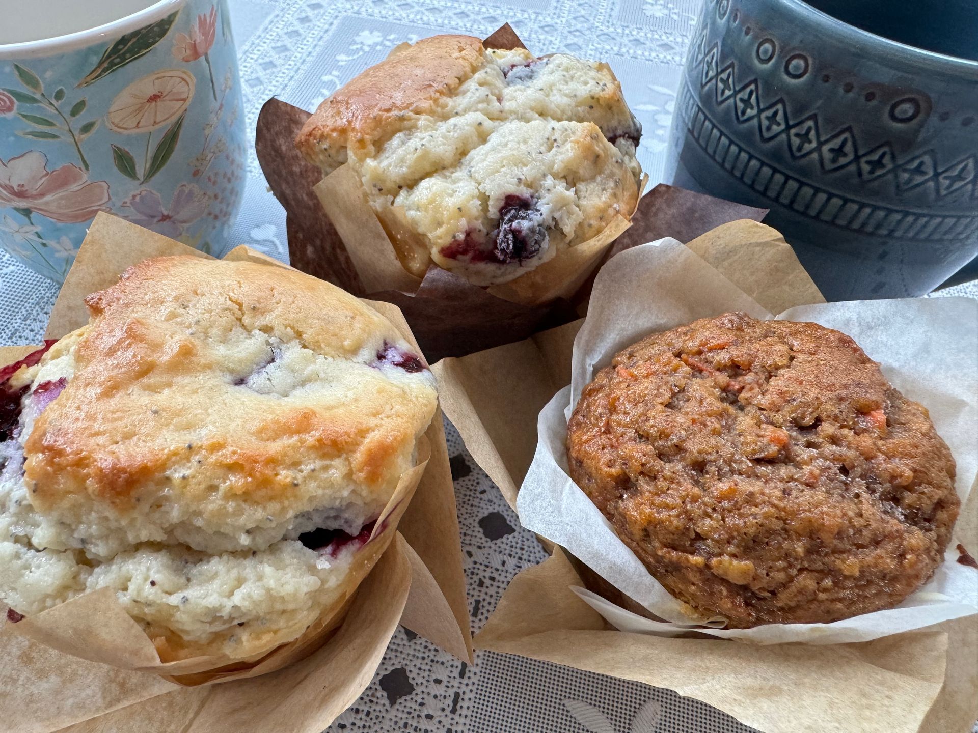 Three muffins—two blueberry and one brown, textured muffin—arranged near two ceramic mugs on a patterned tablecloth.