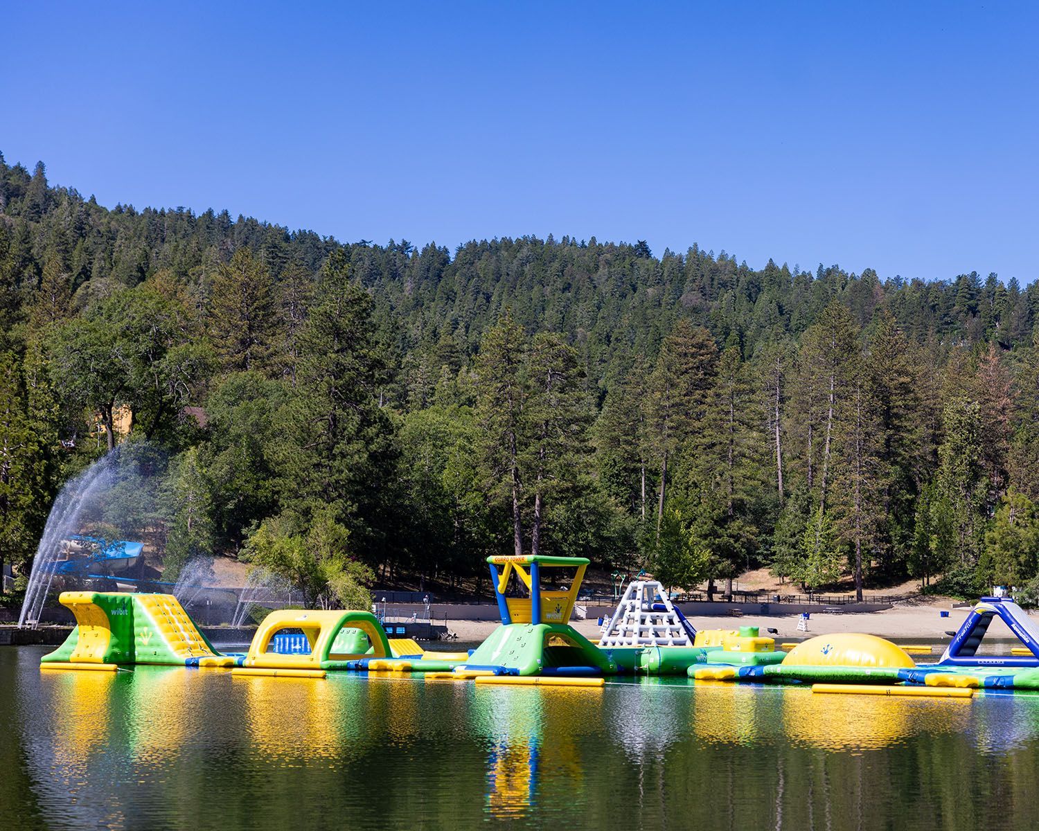 Floating yellow, blue, and green inflatable water park obstacle course on a lake with a forest-covered mountain background.